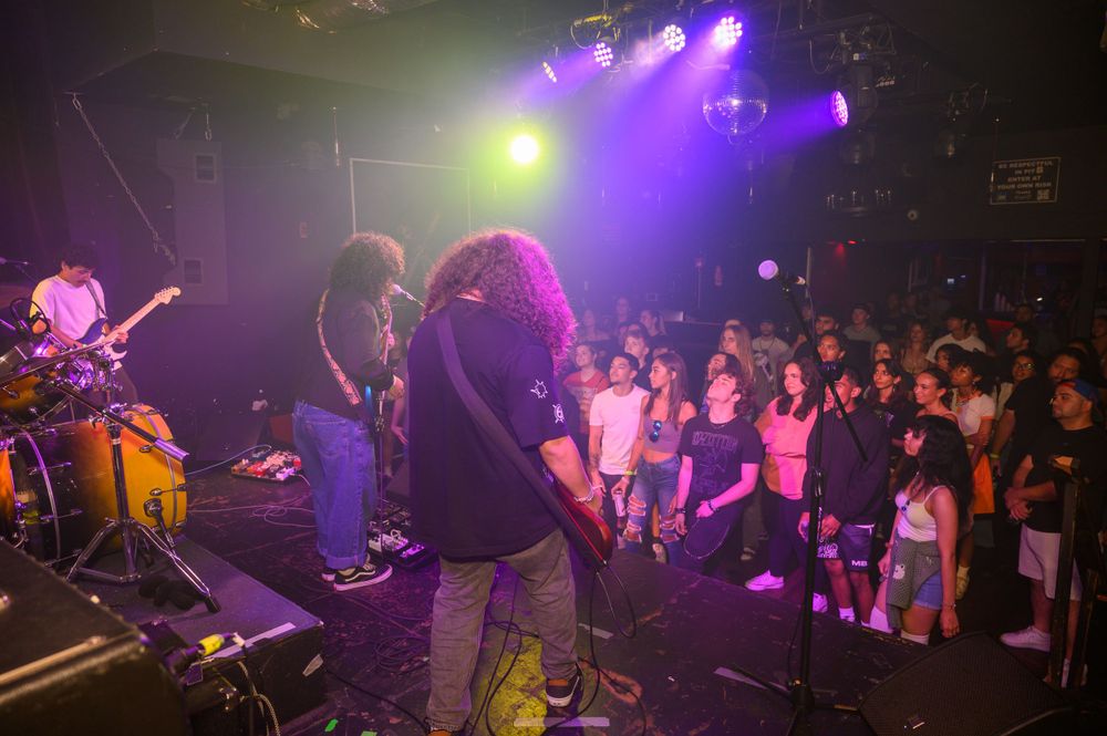 Rock band performs on stage with colorful purple and yellow lighting while crowd watches from floor level.
