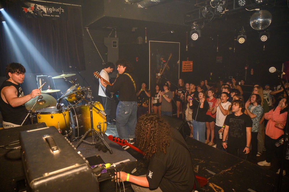 Live concert scene with band performing on elevated stage as audience gathers below in dark venue.