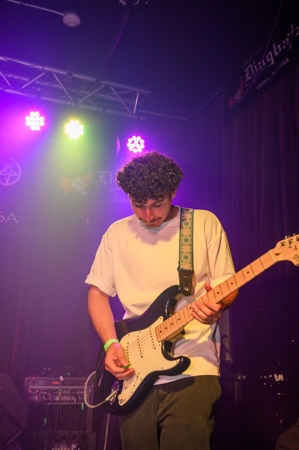 Guitarist wearing white t-shirt plays electric guitar under purple stage lighting at a concert venue.