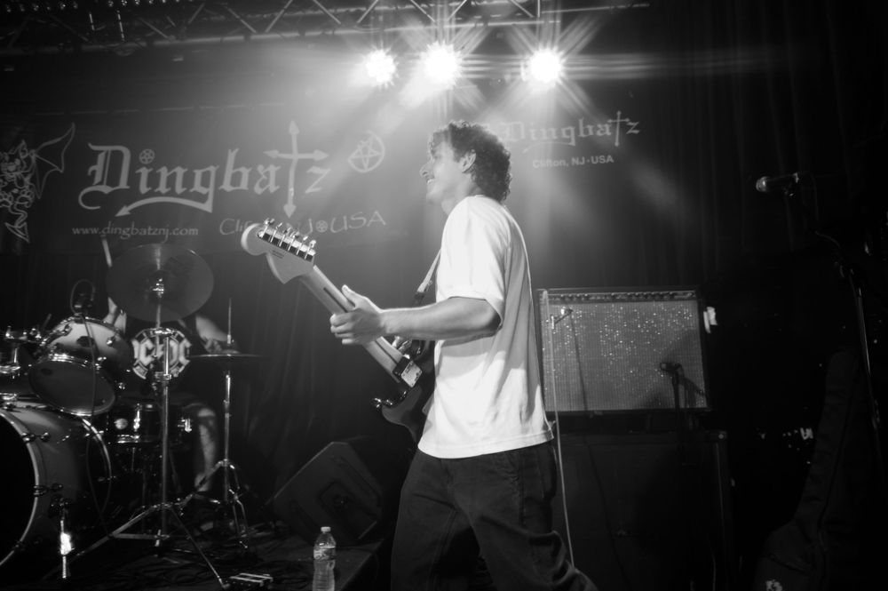 Black and white photo of a performer playing bass guitar on stage at a rock concert with bright stage lights.
