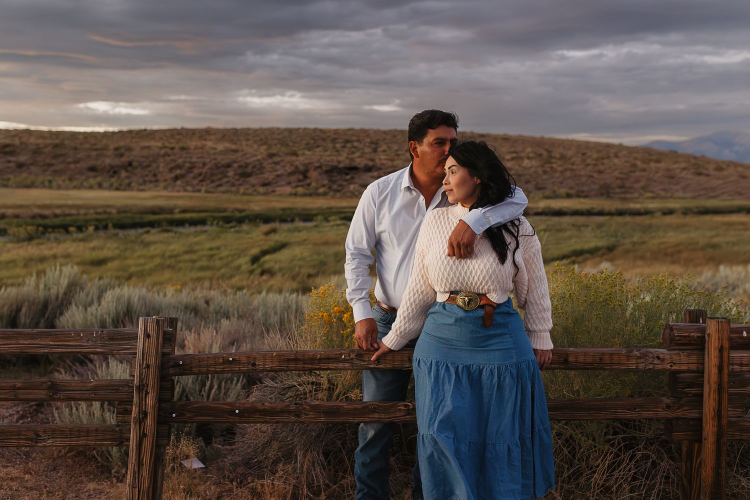Romantic couple poses together against dramatic cloudy skies in an open desert landscape.