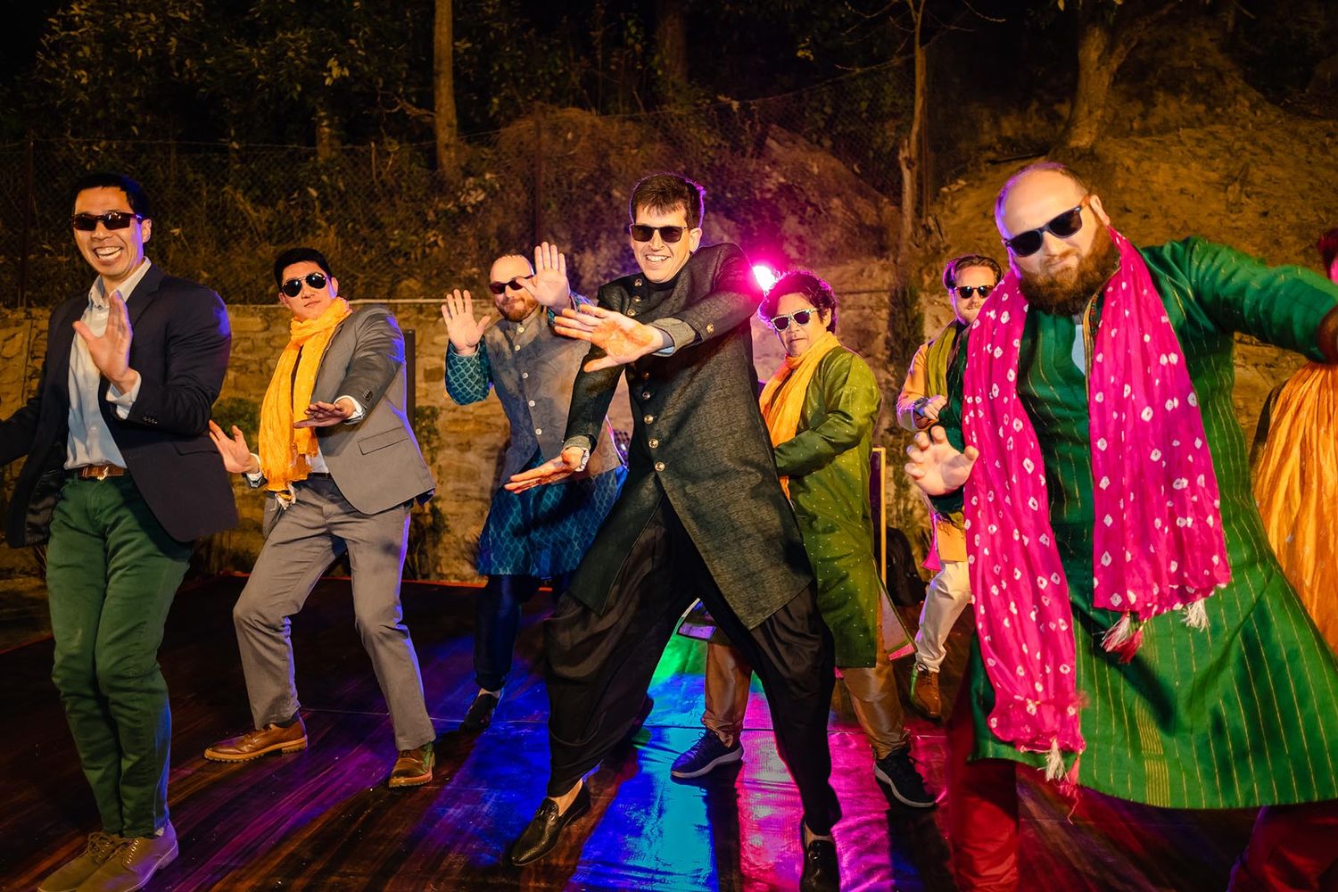 A group of performers in colorful outfits strike dance poses under dramatic lighting on an outdoor stage at night.