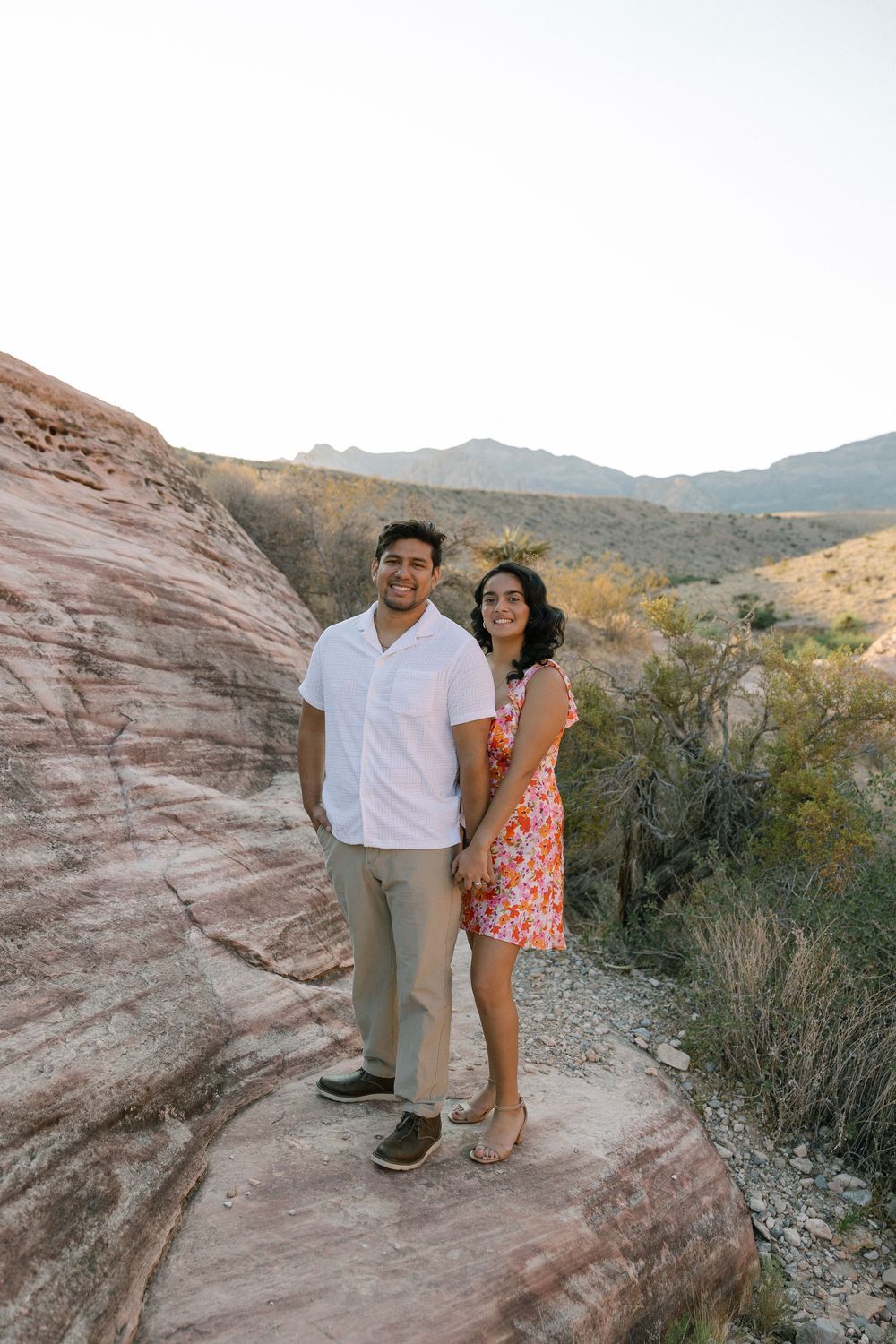 A couple poses together on red rock formations in a desert landscape during sunset.
