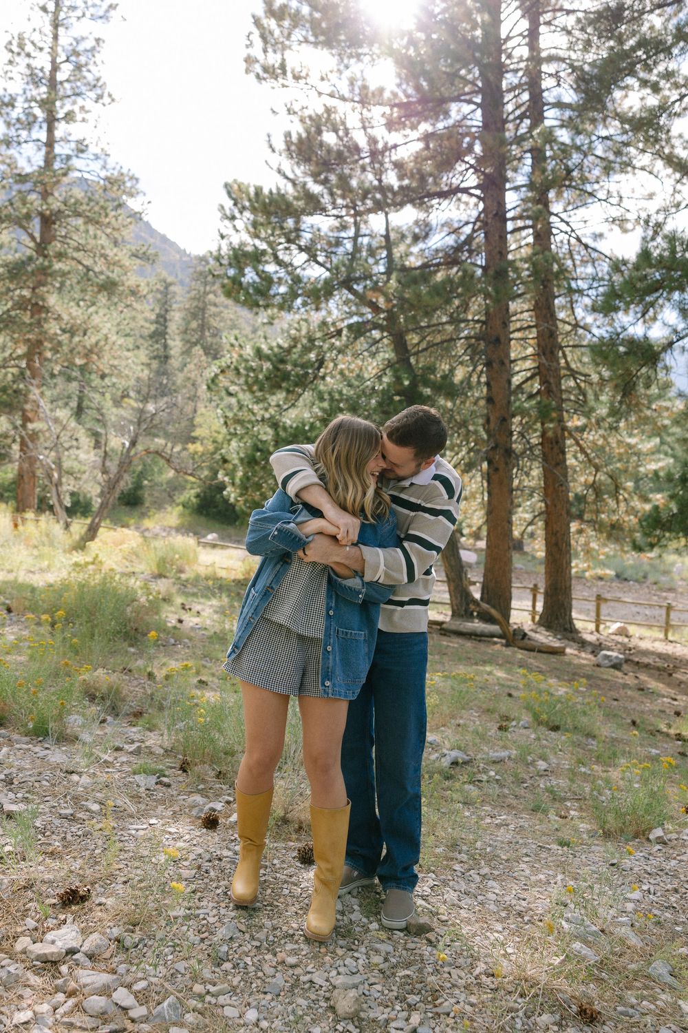 Two people share an intimate moment while hiking in a sunlit pine forest with rocky terrain.