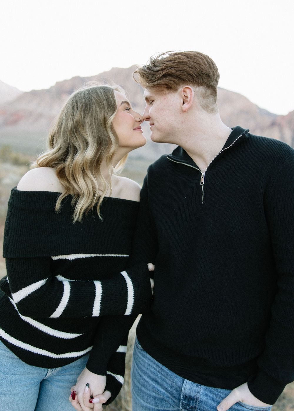 A couple in coordinating black sweaters shares a tender moment against mountain backdrop.