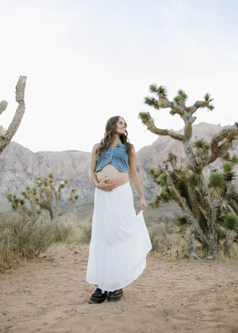 Expectant mother in white dress and denim top poses against desert landscape with Joshua trees.