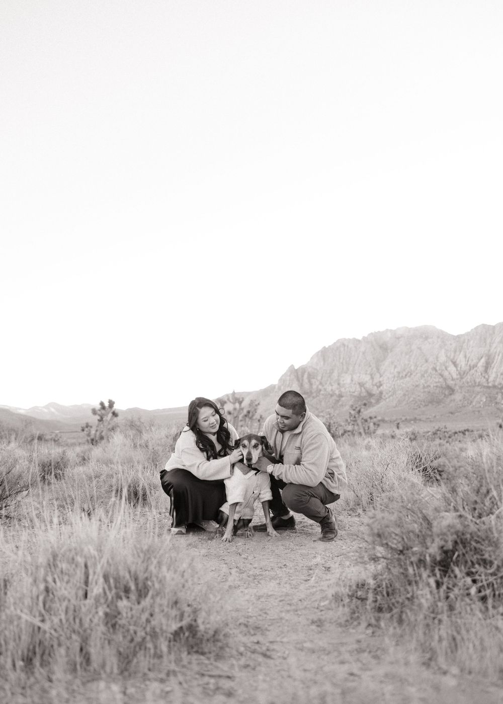 A couple kneels with their dog on a desert trail with mountains in background.