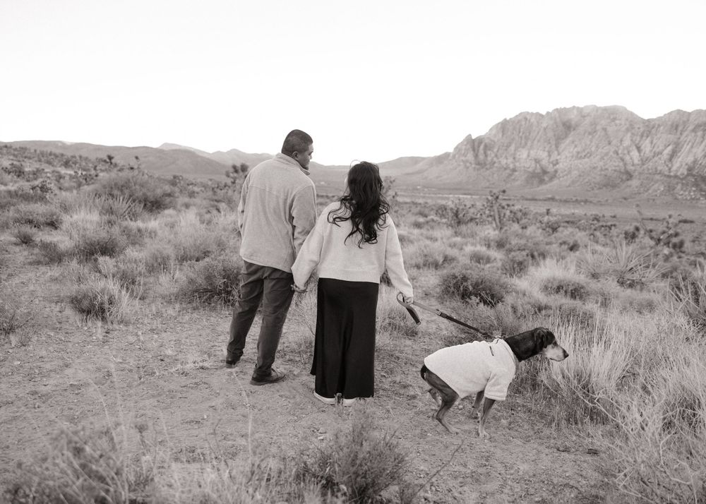 Couple walking with their dog through desert brush with mountains in background, shot in black and white.