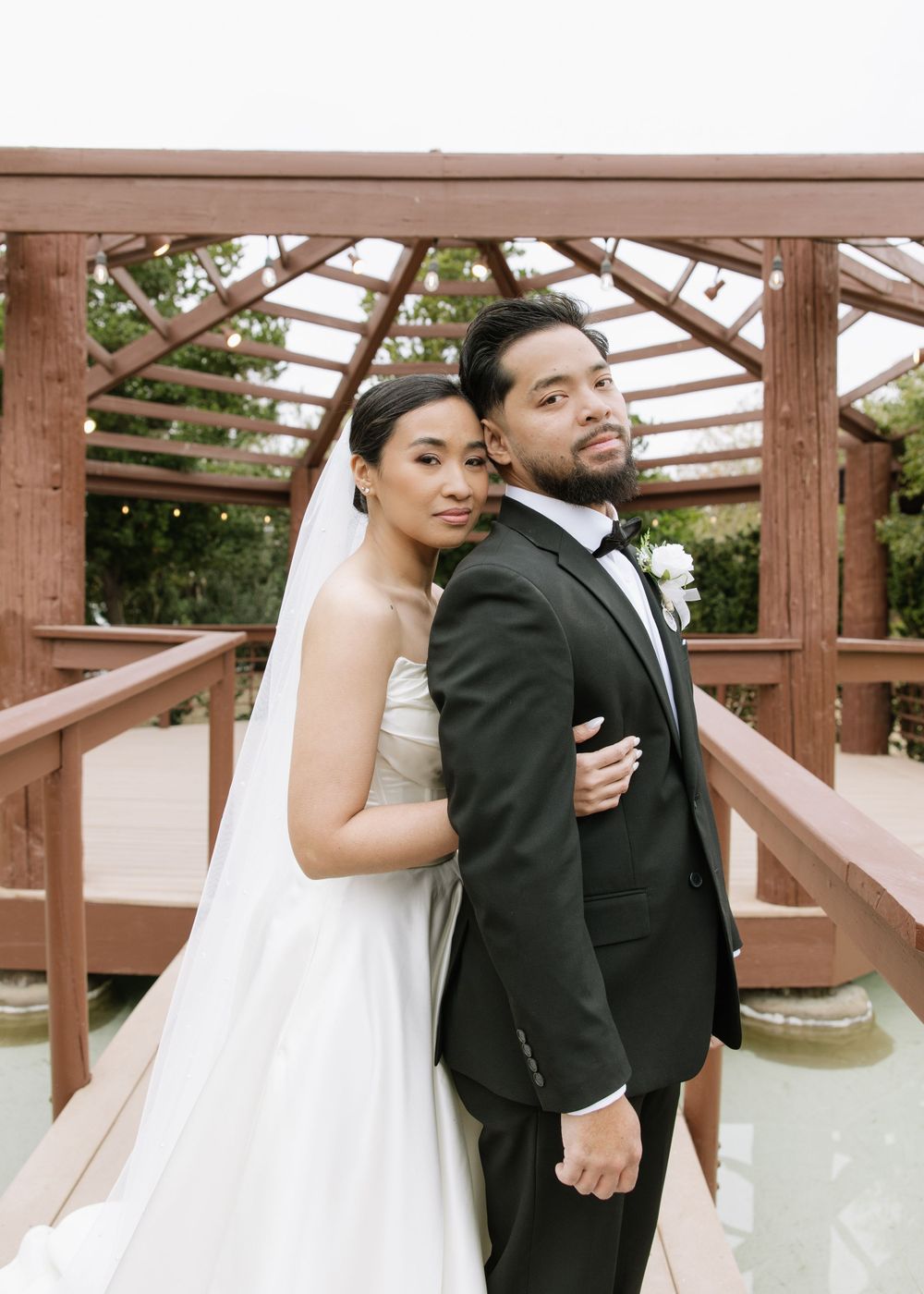 Newlywed couple poses together on wooden bridge gazebo in garden setting.