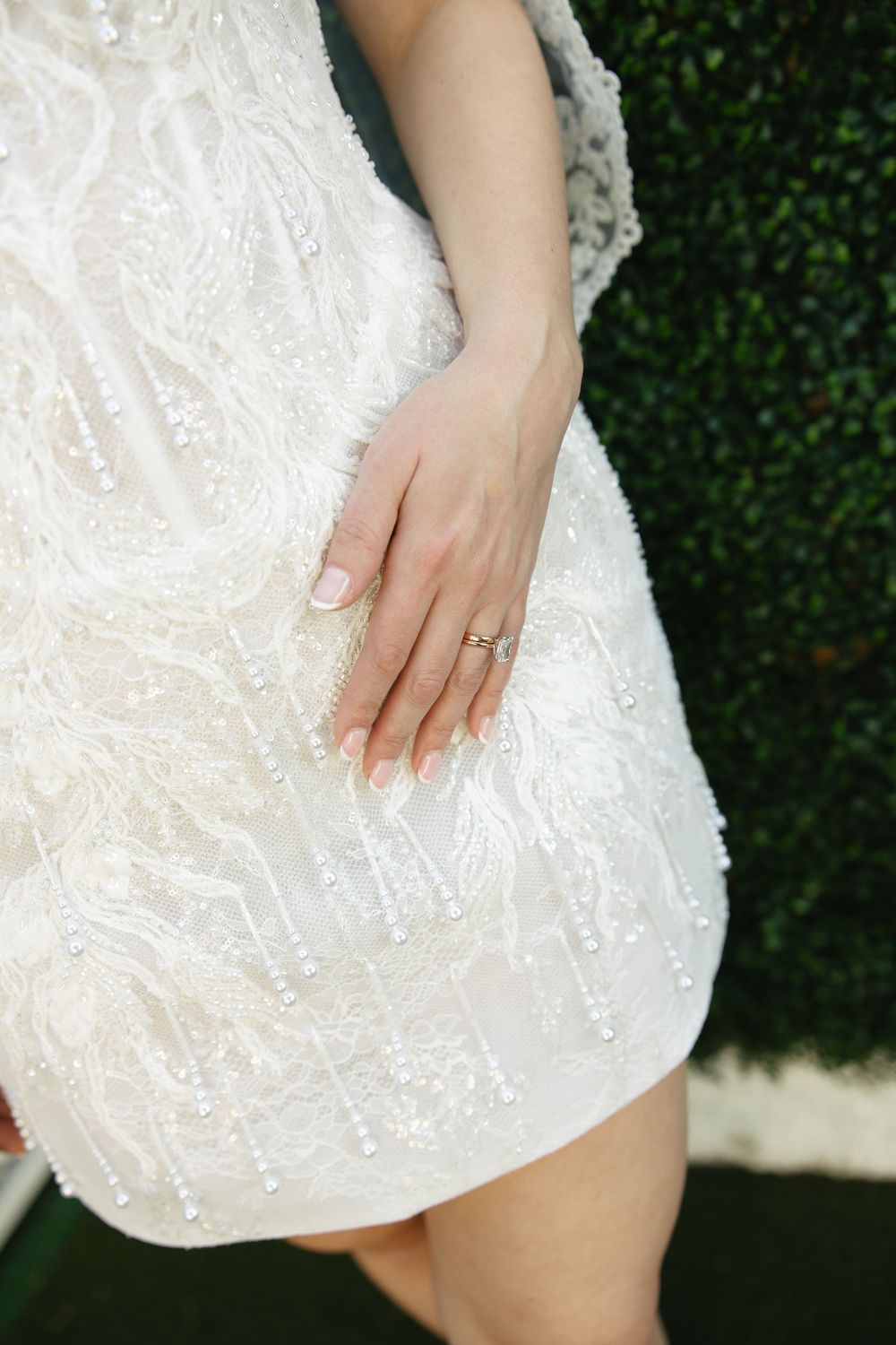 Close-up view of hands resting on white sequined fabric against a green hedge background.