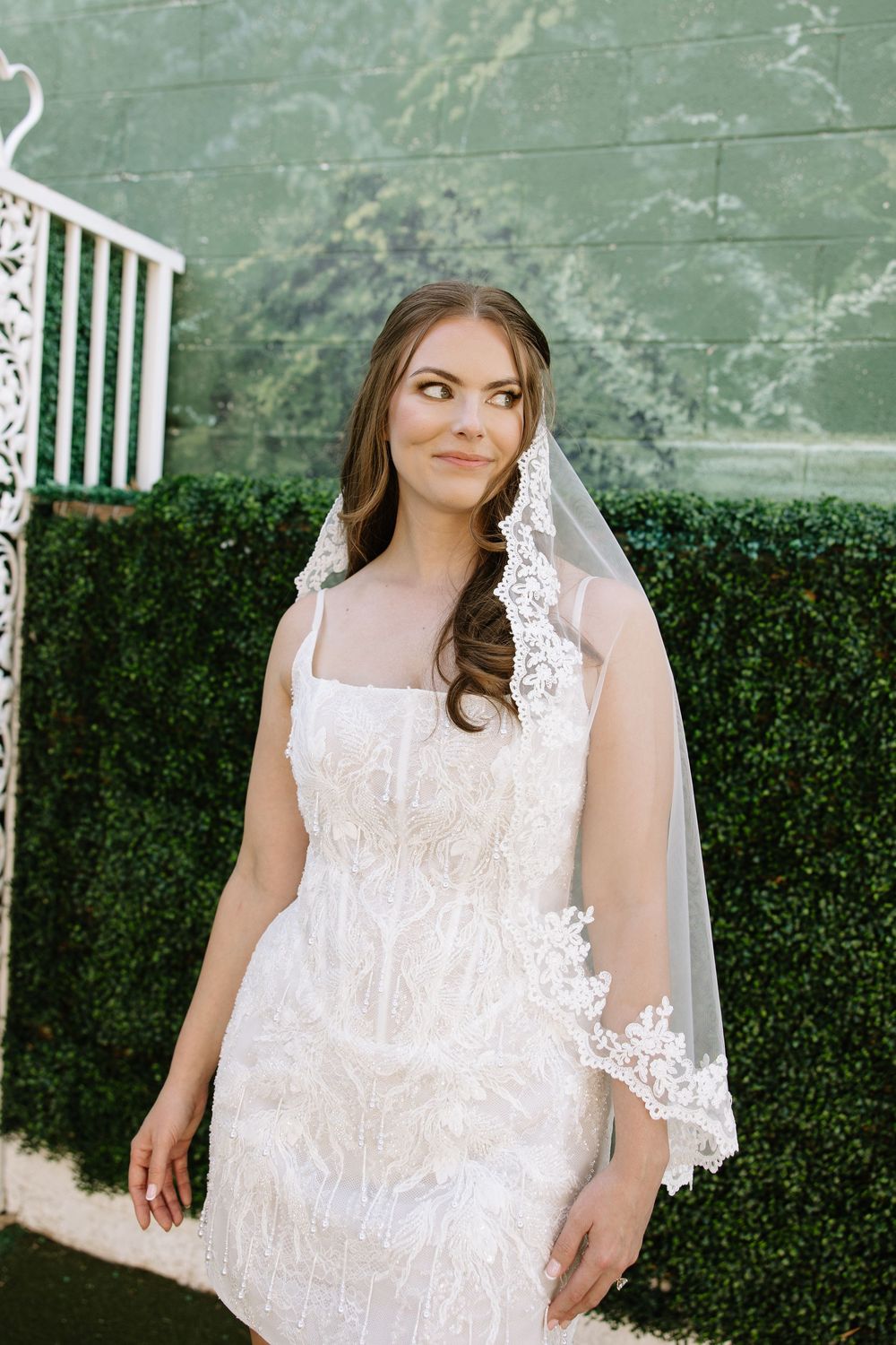 A bride in a white lace wedding dress and veil stands in front of a green hedge wall outdoors.
