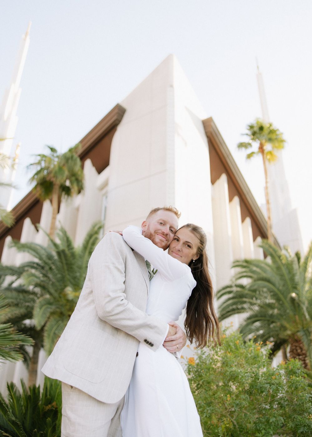 Couple embraces outside modern white church with palm trees.