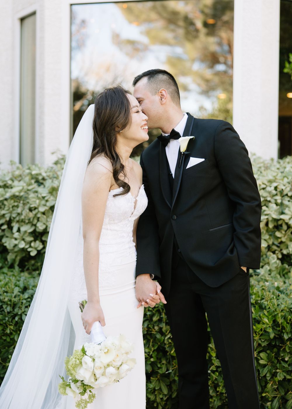 Romantic outdoor wedding portrait with white bouquet and long veil against garden backdrop.