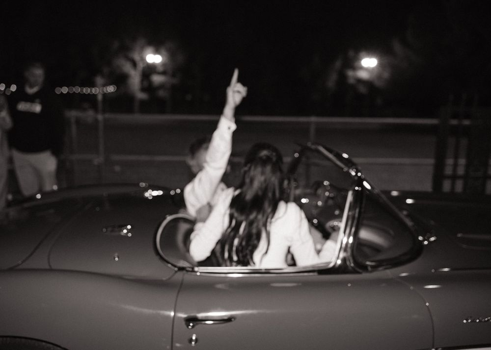 Black and white photo of someone in a vintage convertible car at night pointing upward.