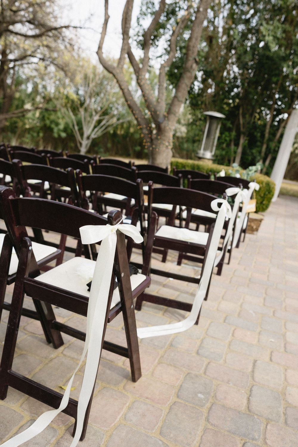 Row of dark wooden folding chairs with white ribbons set up for outdoor ceremony.