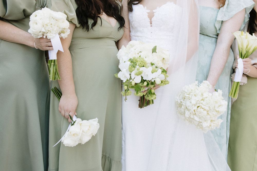 Bridesmaids in sage green dresses holding white floral bouquets next to bride in white gown.