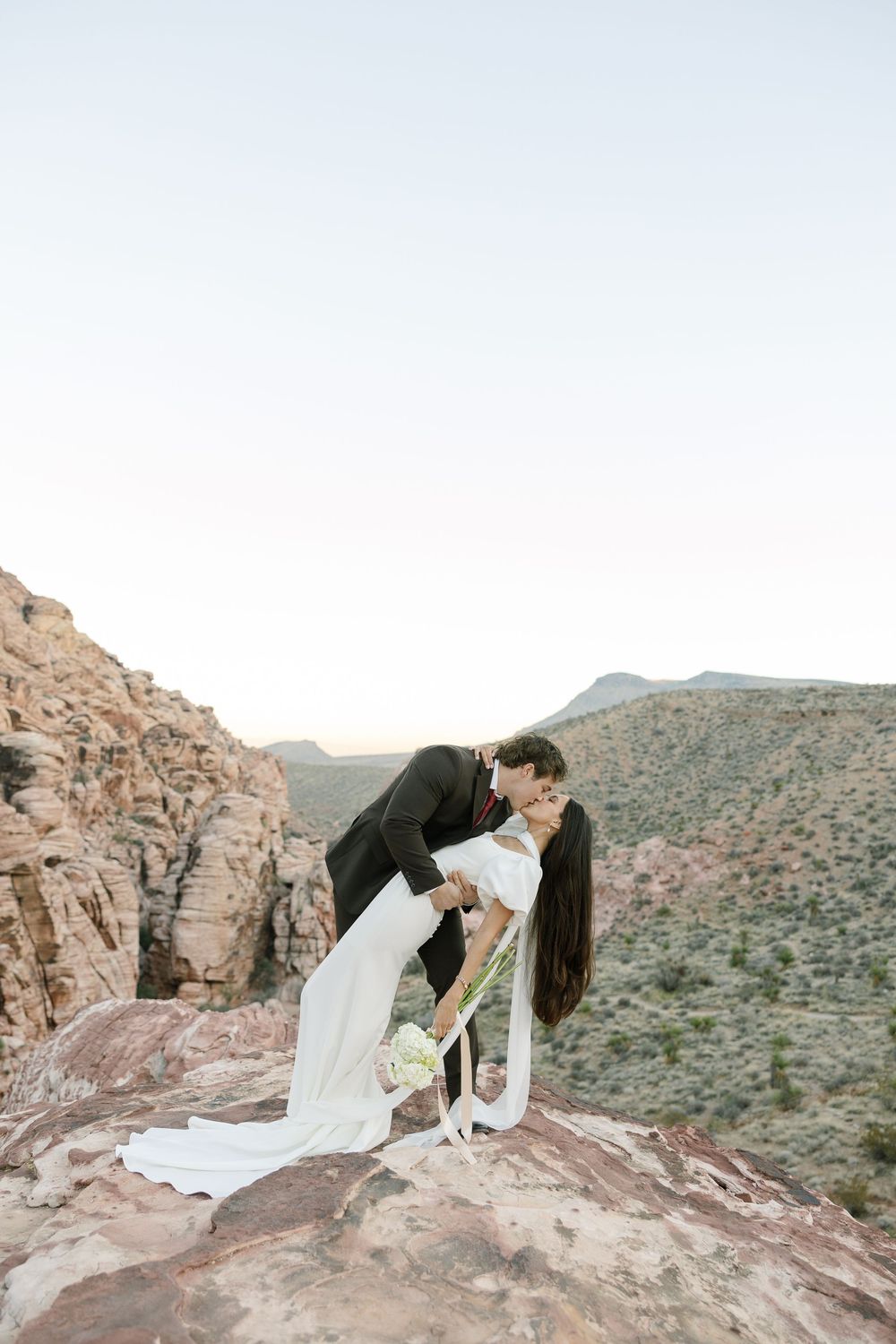 A couple in wedding attire shares romantic moments on red rock formations in a desert mountain landscape.
