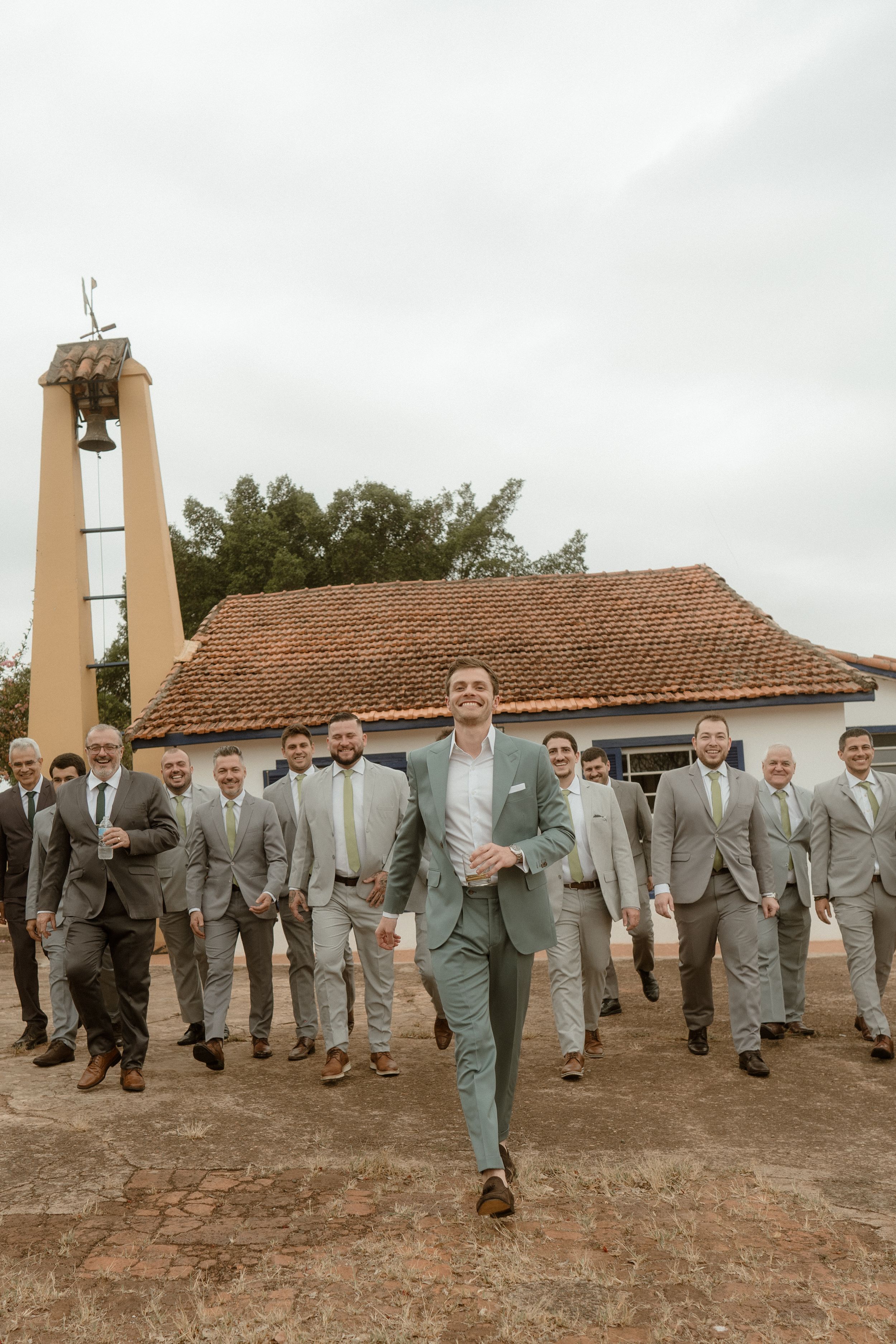 A wedding party in gray suits gathers outside a rustic church with a bell tower.
