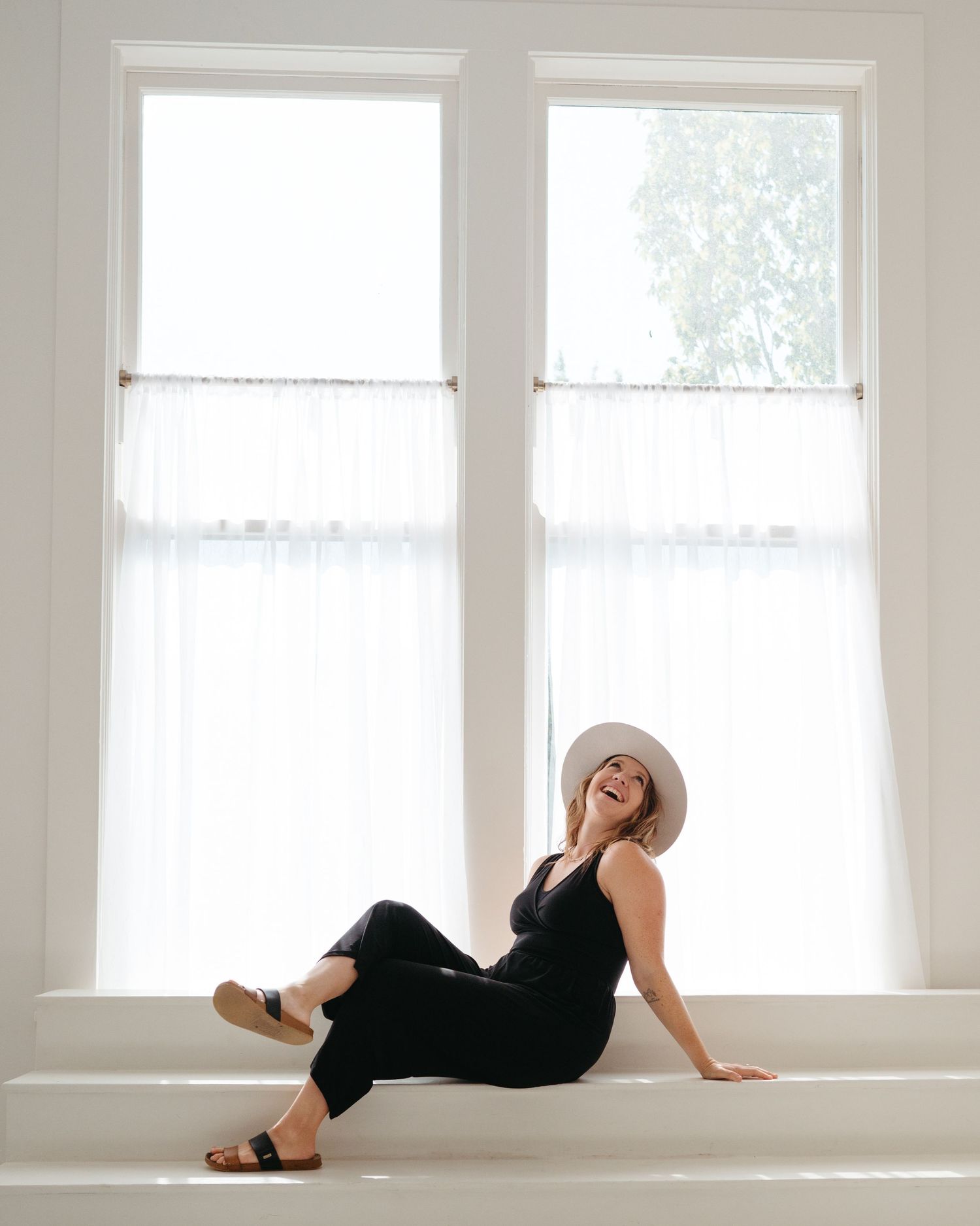 Person in black outfit lounging by bright windows with white curtains.