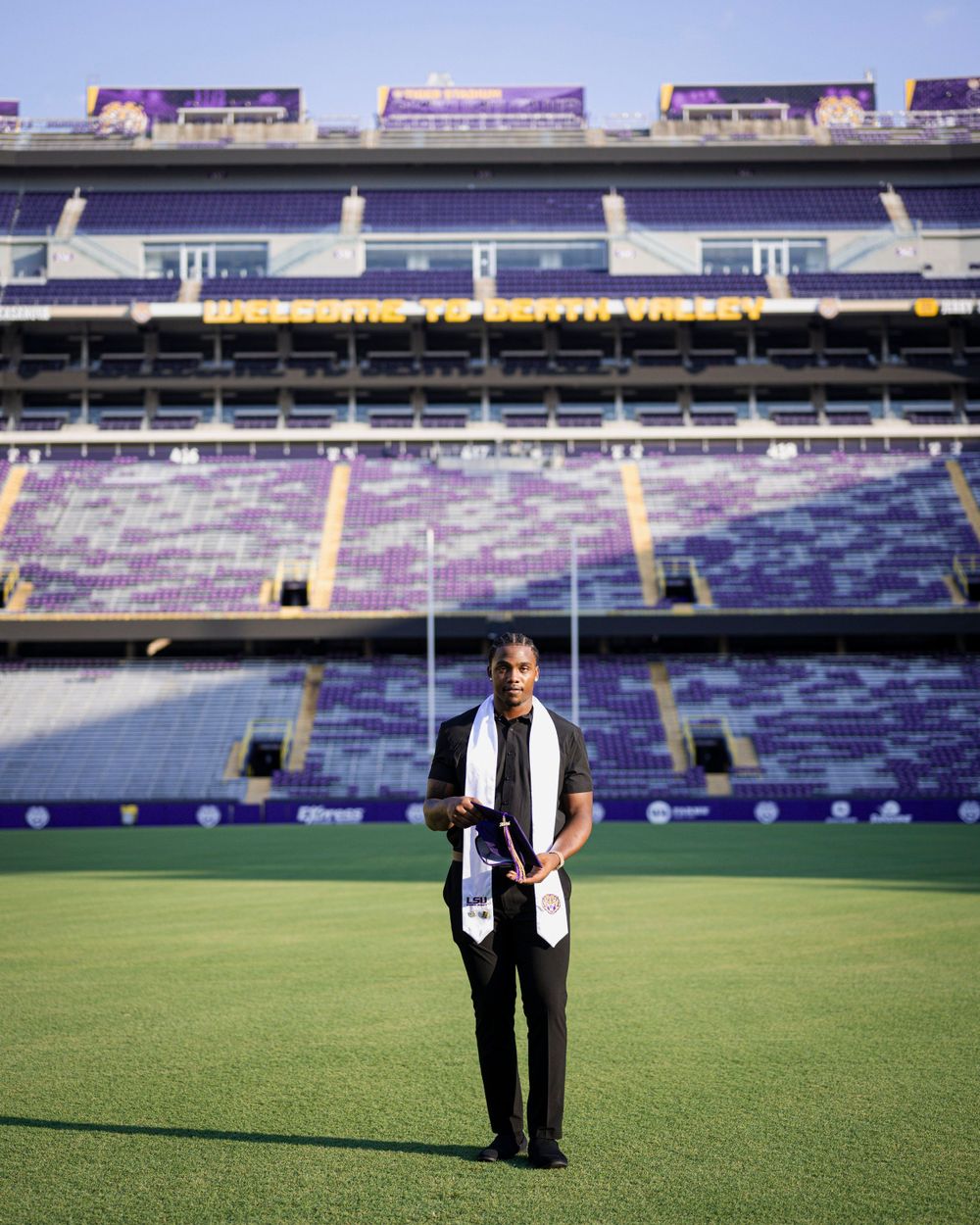 Graduate standing in formal attire on a football field with stadium seating in the background.