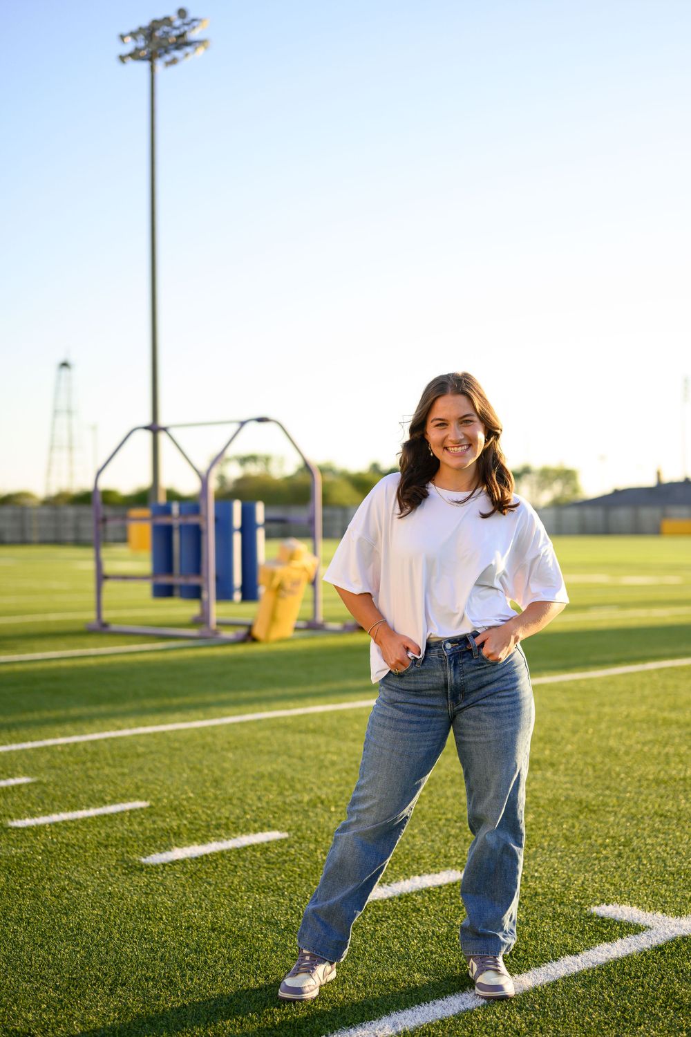 Athletic figure in a white t-shirt and jeans stands confidently on football field lines with stadium lights behind.