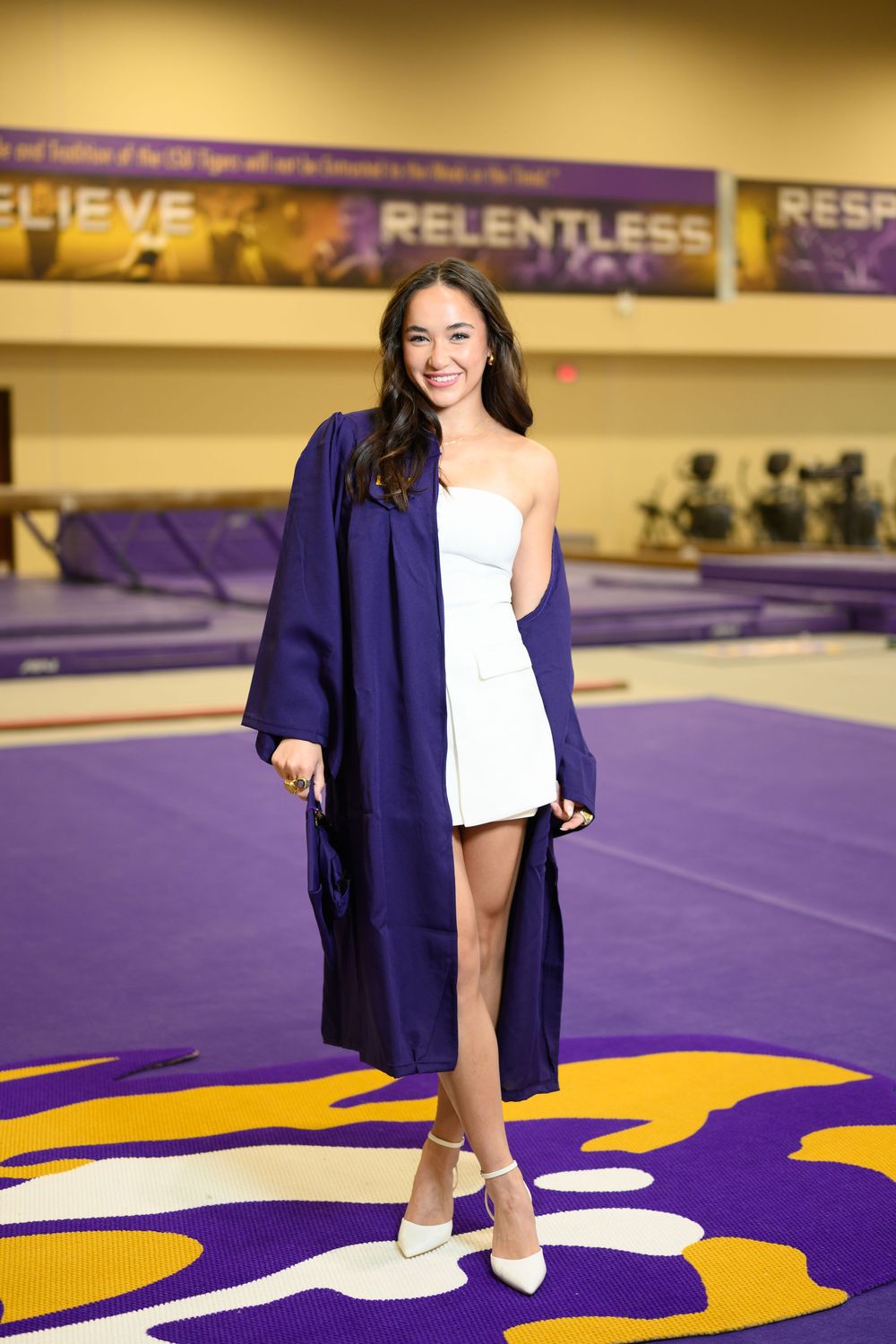 A graduate in white dress and navy graduation gown poses on a purple and gold gymnasium floor with banners overhead.