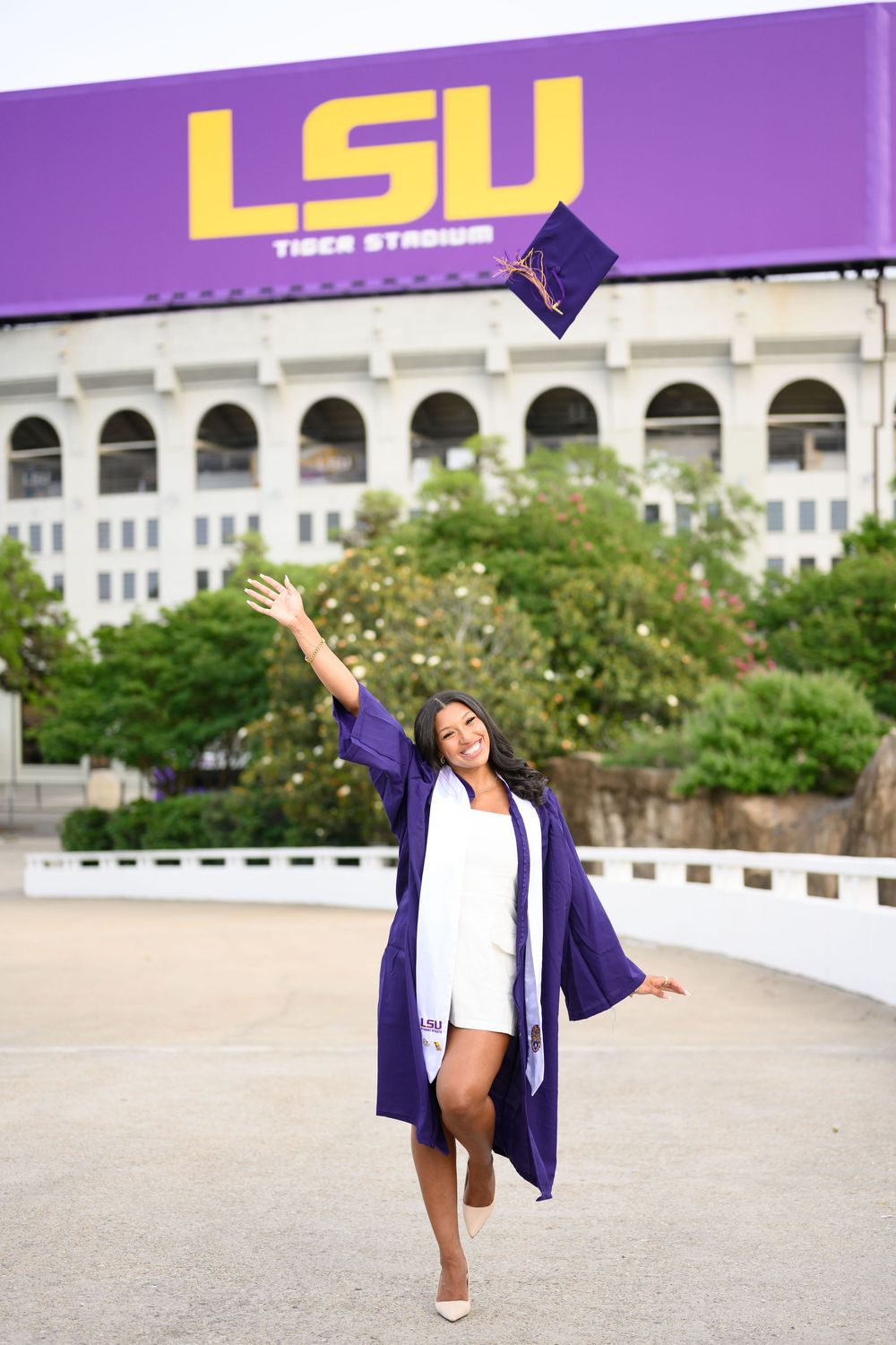 A graduate poses joyfully in front of LSU Tiger Stadium, tossing their graduation cap in celebration.