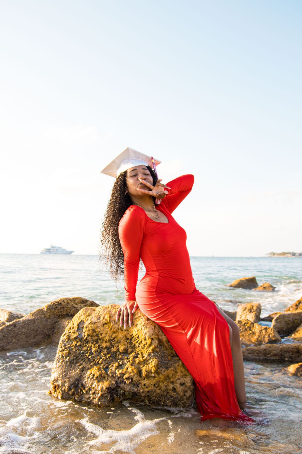 A person in a flowing red dress poses on coastal rocks at sunset by the ocean.