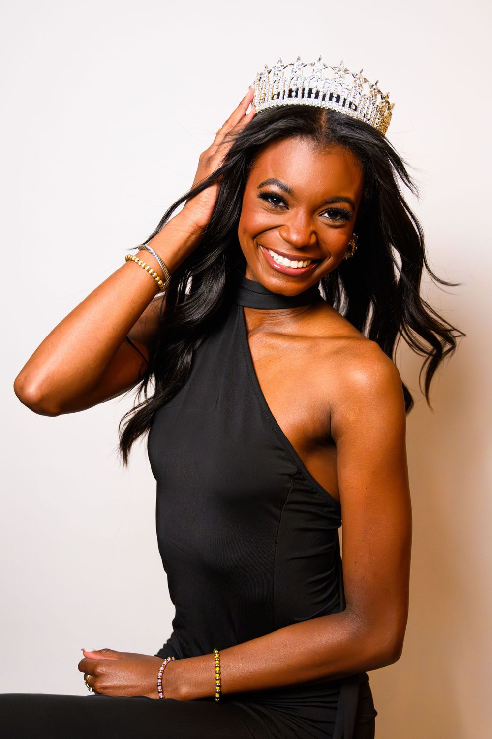 Pageant winner in black one-shoulder gown wearing crystal crown and radiant smile.