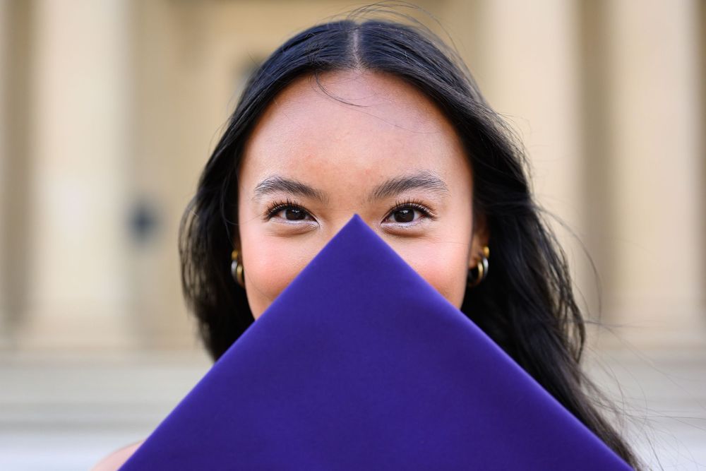 A person holds up a bright purple folder or book, partially covering their smile as they peek over the top.