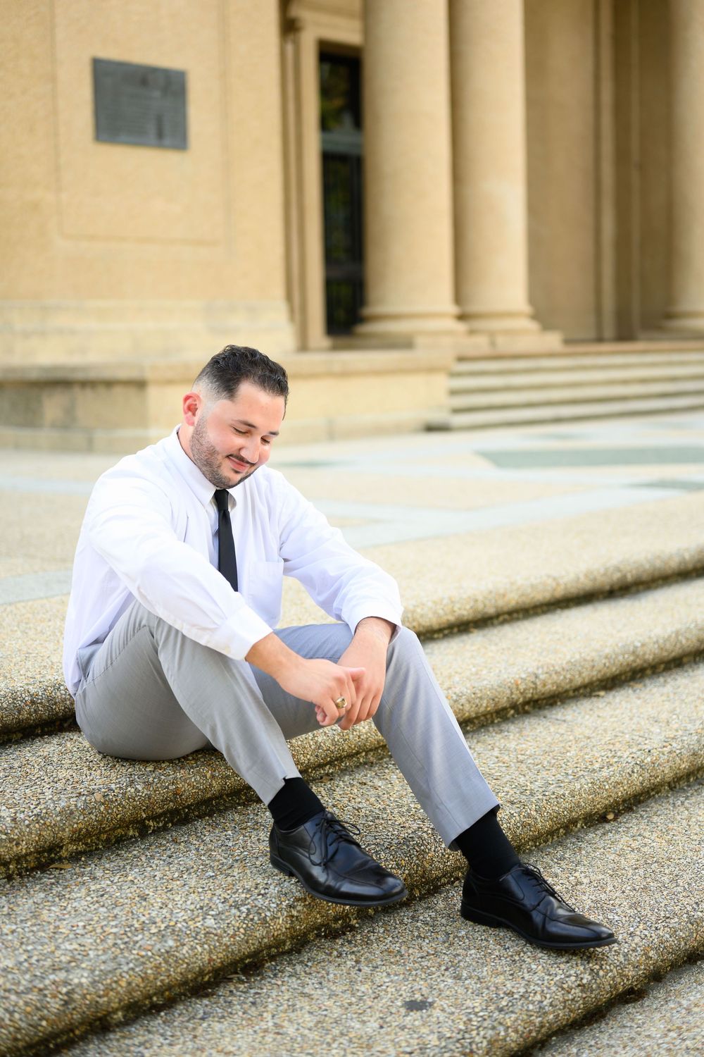 Professional in business attire sitting thoughtfully on concrete steps outside a building with classical columns.