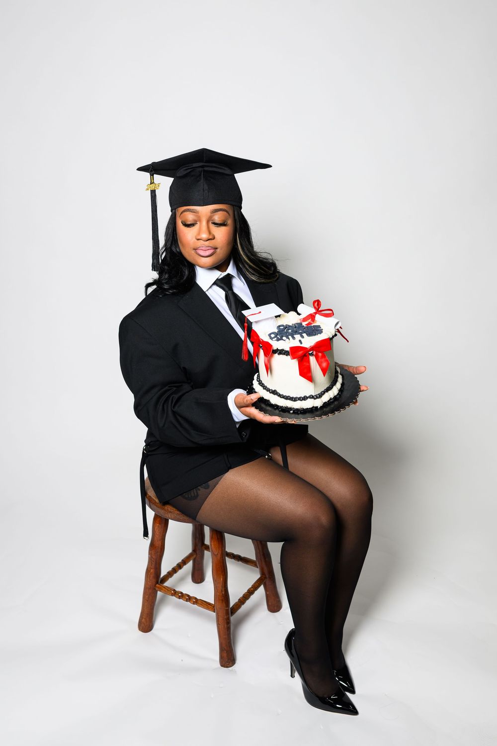 Graduate in cap and gown sits on wooden stool holding decorated cake in celebratory graduation photo series.