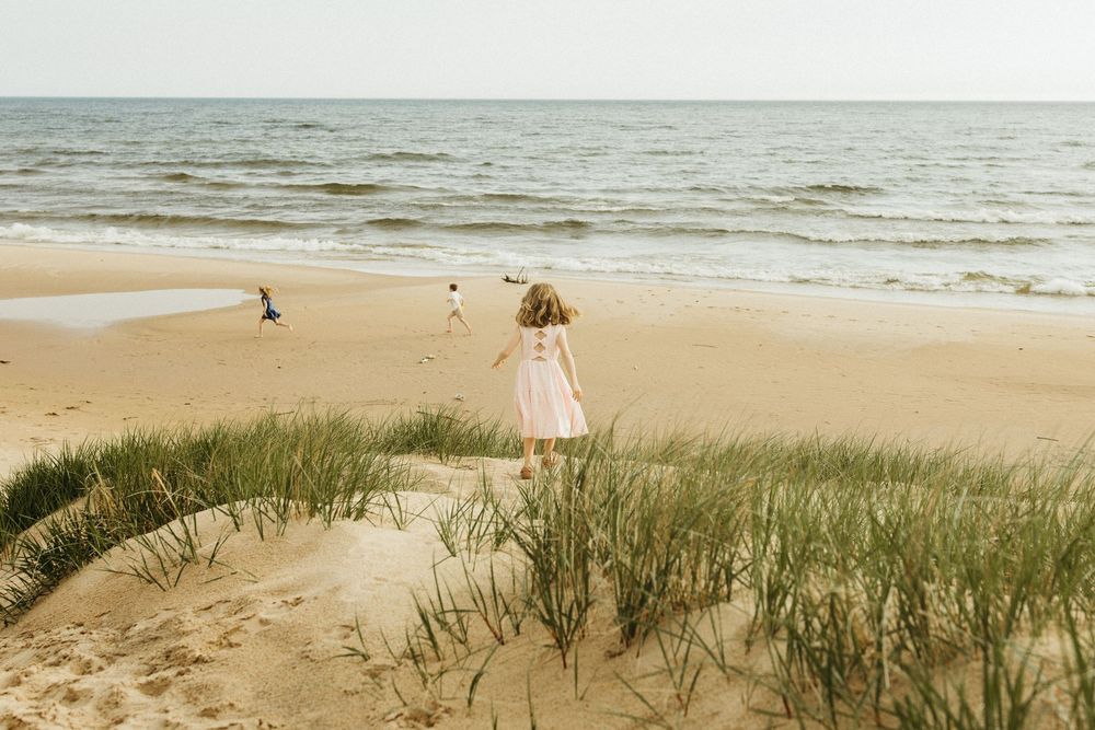 Young girl in white dress walking on sandy beach toward ocean waves with grass dunes in foreground.