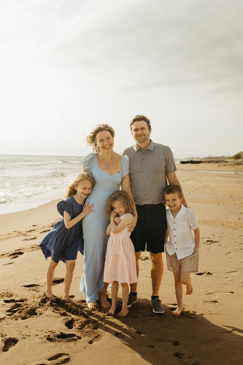 Family of five smiling together on a sandy beach at sunset, with ocean waves in the background.