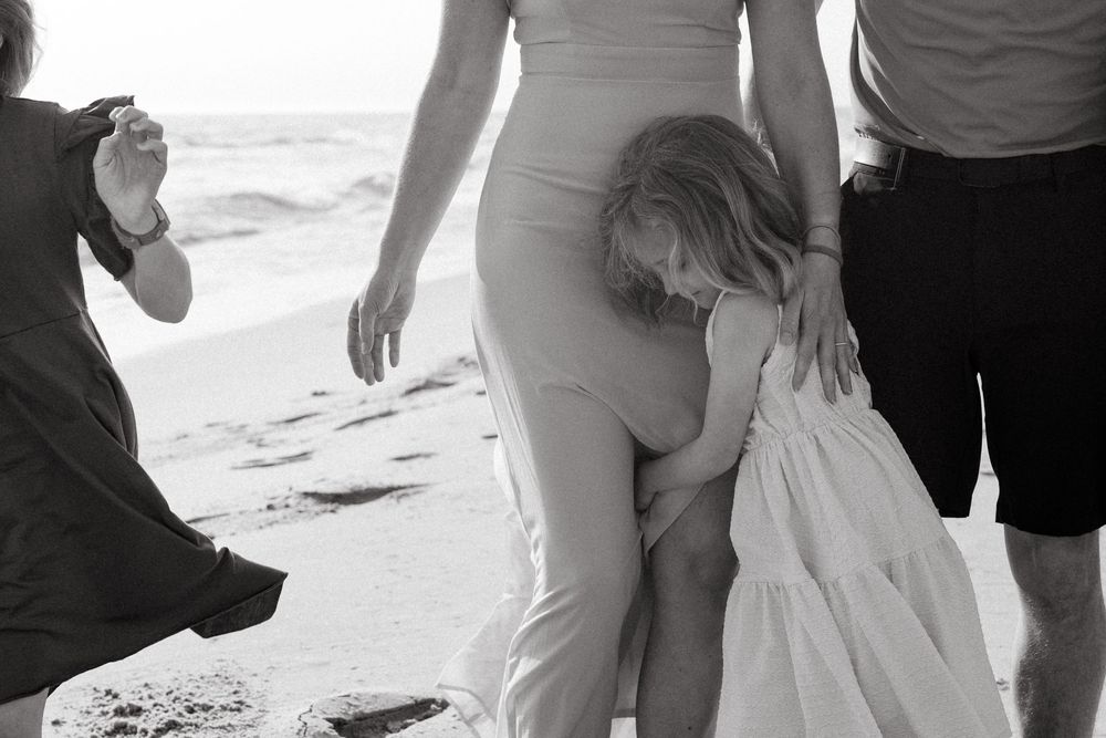 Black and white beach photo of a child hugging a woman in a white dress while others gather nearby on sandy shore.