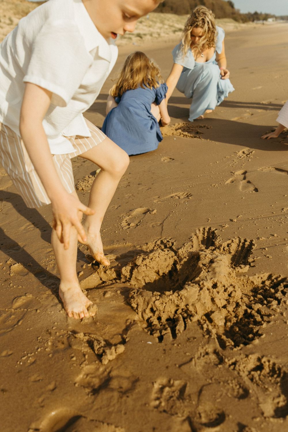 Family playing in the sand on a sunny beach, with children and a woman crouching near a small sandcastle.
