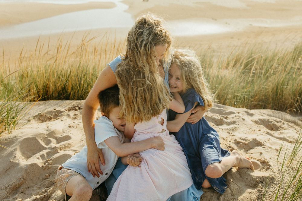 A mother hugs her three children on sandy dunes with golden grass in the background.