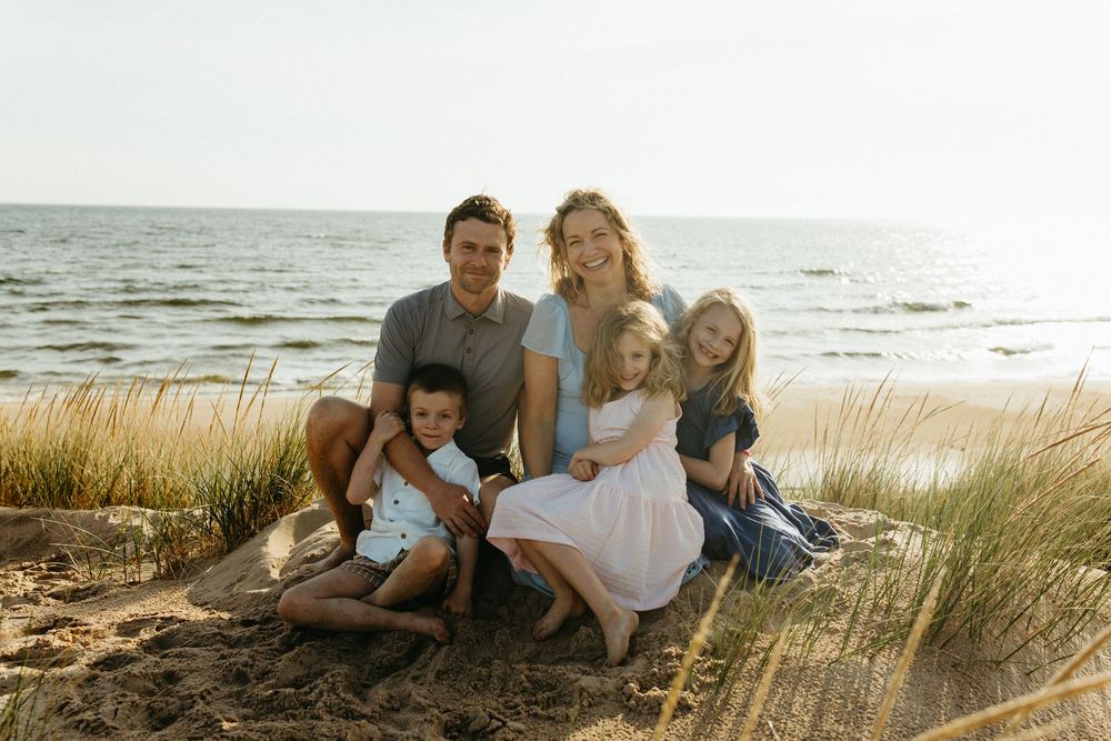 Family of five posing together on sandy beach dunes with ocean in background on a sunny day.