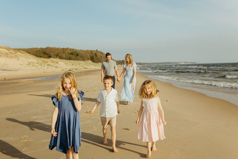 Family of five walking on a sunny beach, two parents and three children in summer dresses enjoying a seaside stroll.
