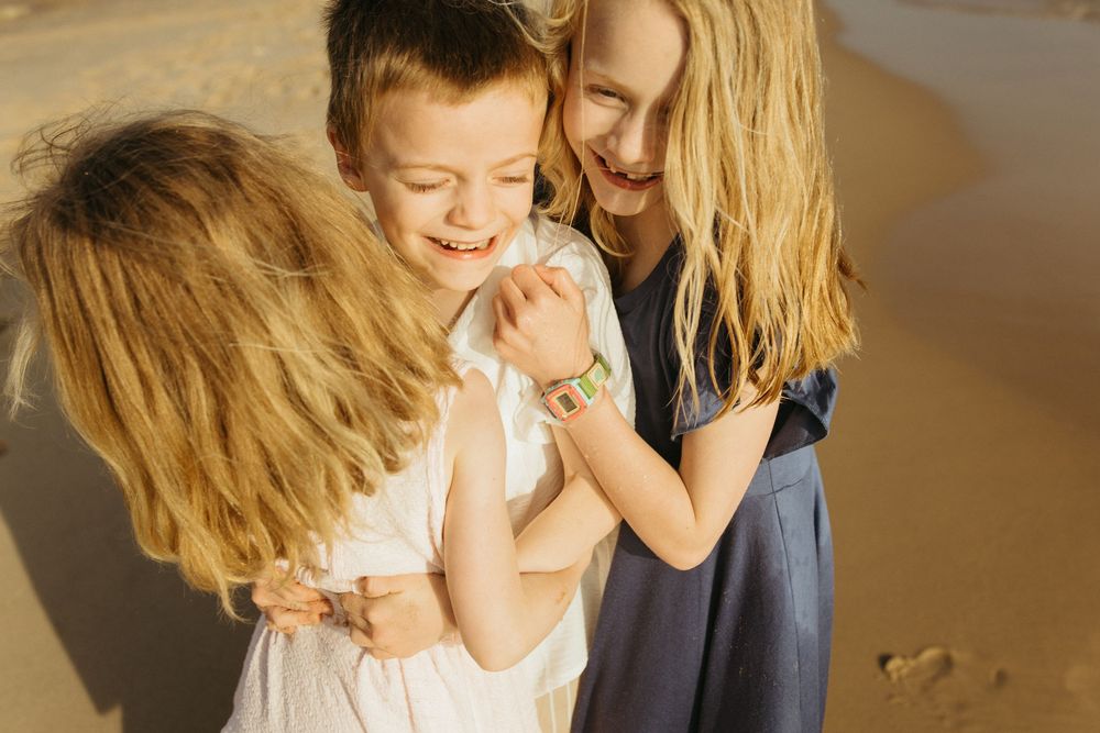 Three happy children hugging and laughing together on a sunny sandy beach.