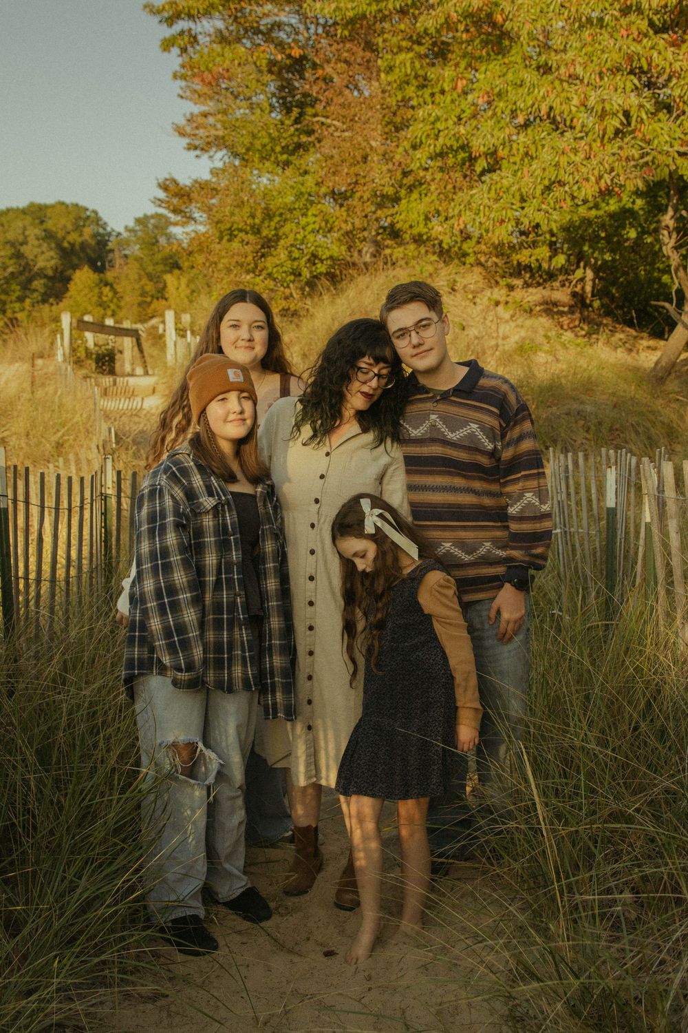 Family of five posing on a sandy beach path surrounded by autumn foliage during golden hour.