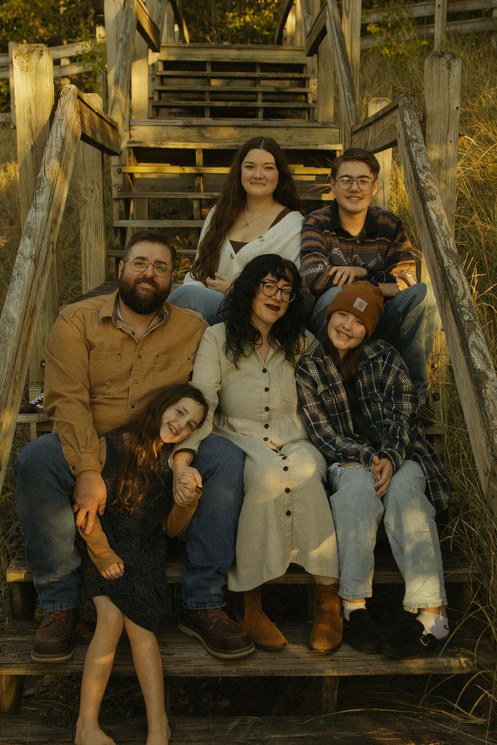 Family of six sitting together on wooden beach stairs smiling during warm sunset light.