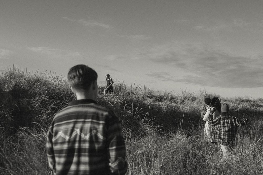 Black and white photo of people walking through tall grass dunes on a beach.