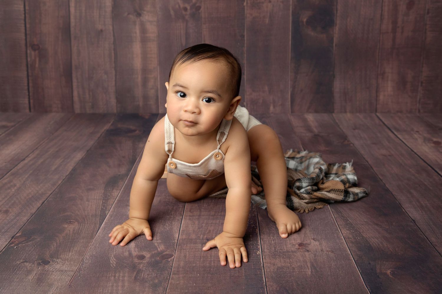 Small infant crawling on wooden floor wearing tan overalls.