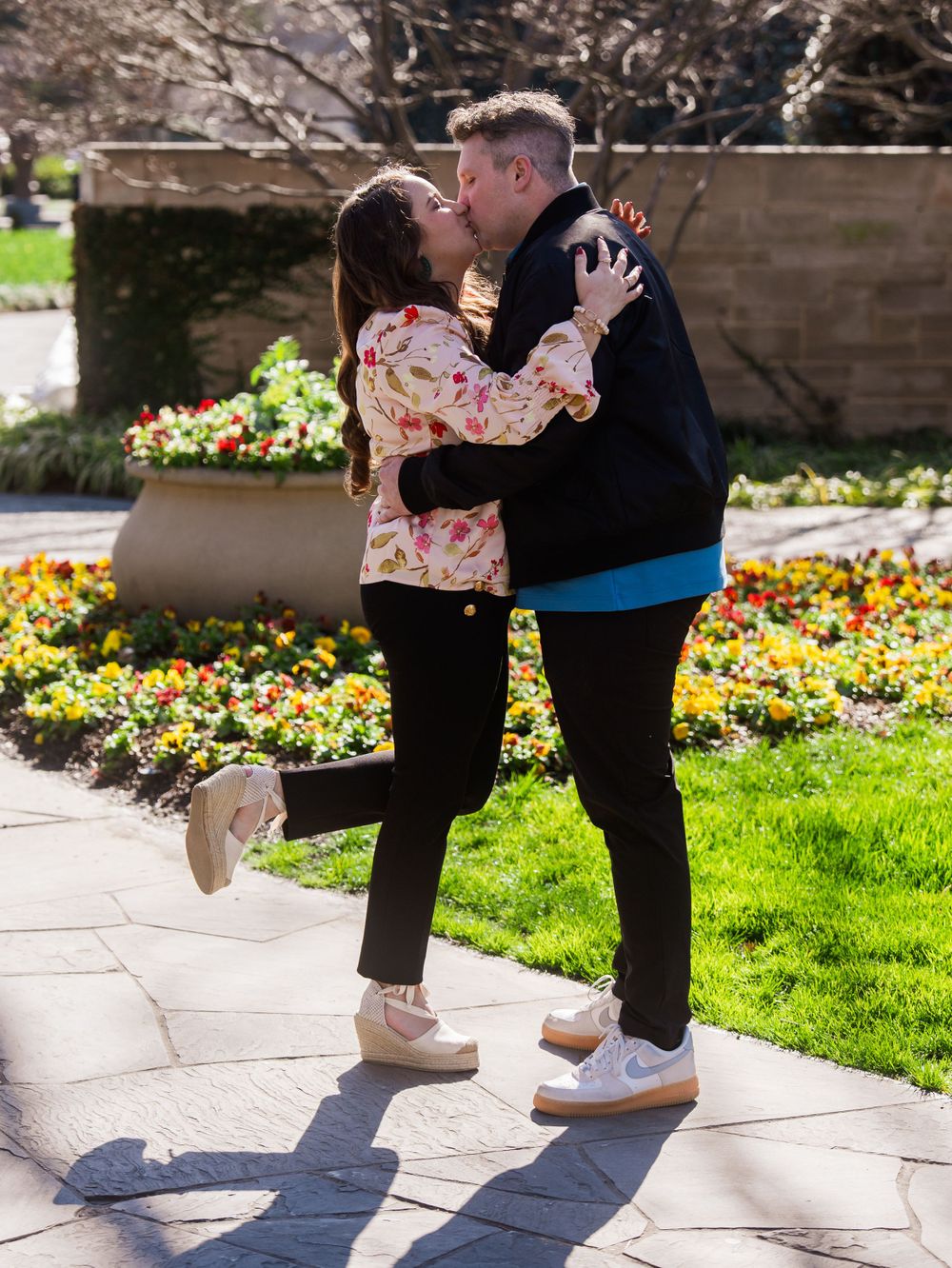 A couple shares a romantic kiss in a spring garden with colorful flowers blooming in planters behind them.
