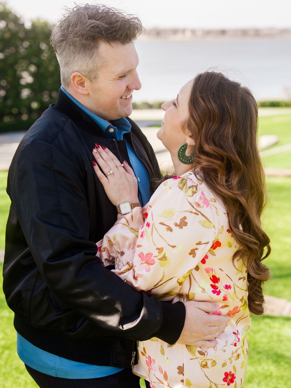A romantic couple embraces outdoors wearing a black jacket and floral dress by the water.