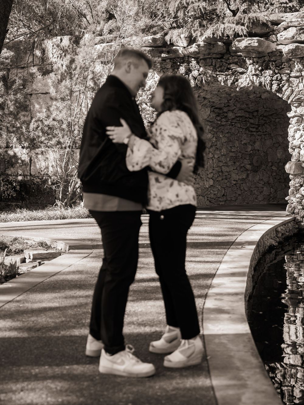 A black and white photo captures a couple in an intimate embrace against a stone wall backdrop.