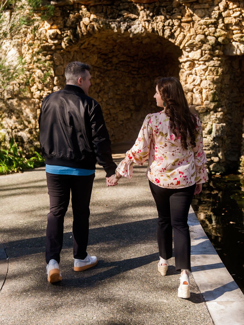 A couple walks hand in hand through a stone archway passage lit by natural sunlight.