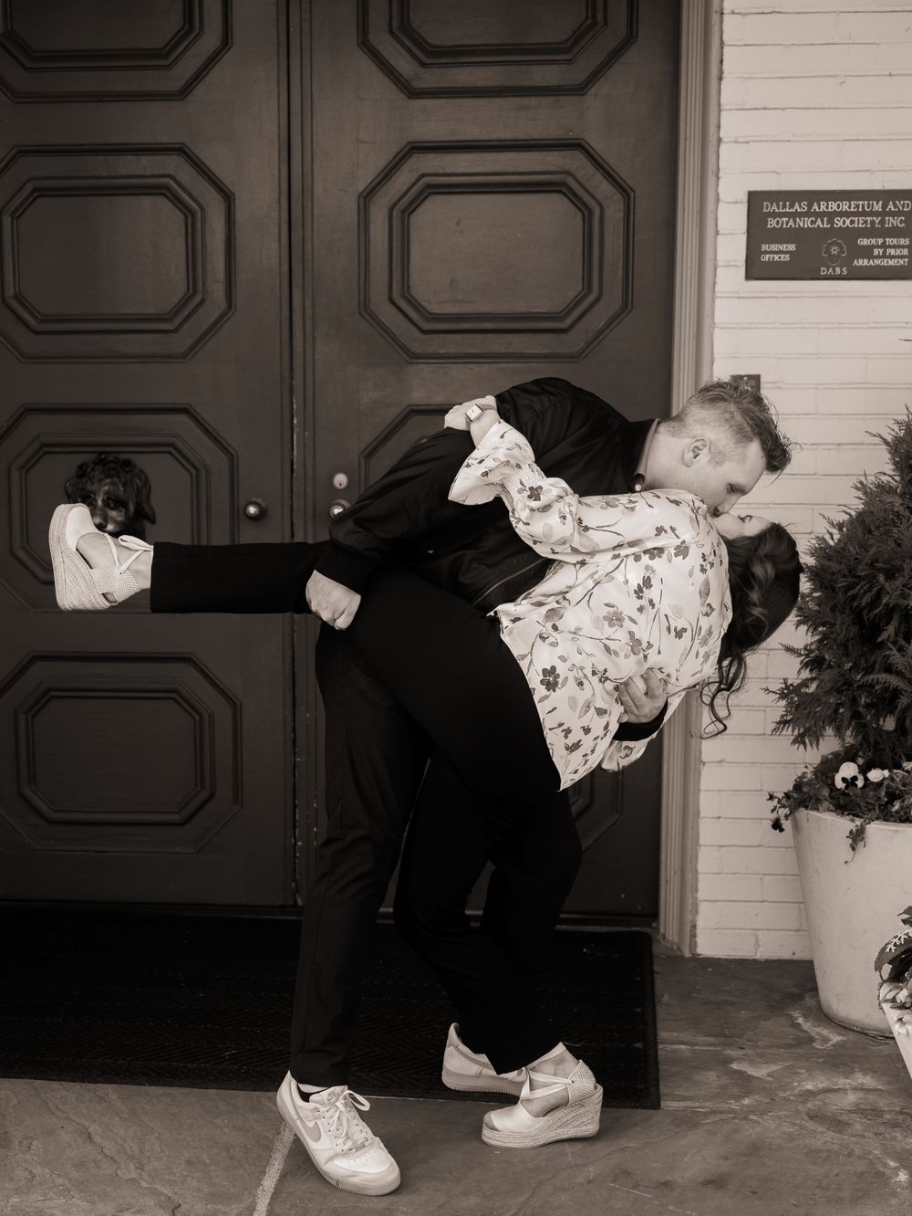 A couple in retro attire share a romantic dip pose outside black double doors in a nostalgic black and white photo.