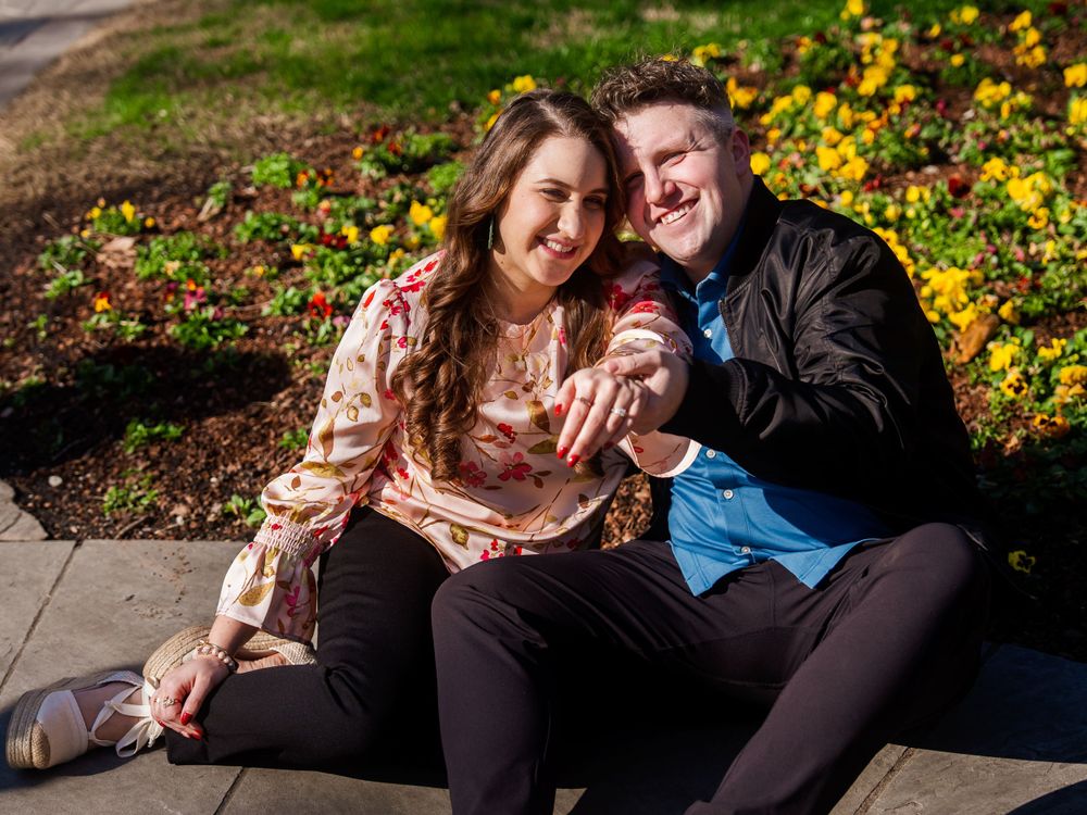 A happy couple sits together outdoors on a sunny day with yellow flowers blooming in the background.